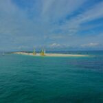 Una lunga lingua di sabbia bianca dove sono tirati a secco catamarani dalle vele gialle e bianche, circondata da barche a vela all'ancora su un'acqua turchese, sotto un cielo blu striato di nuvole.