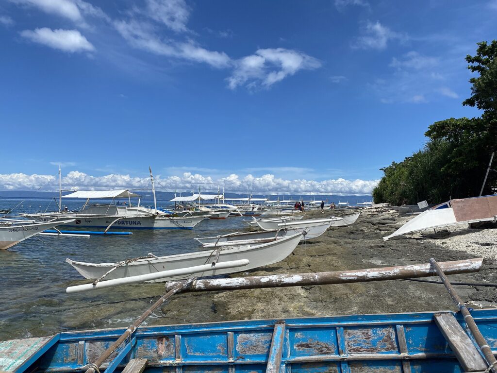 Una fila di bangka bianche ormeggiate lungo una riva rocciosa, piccole piroghe a bilanciere tirate sulla roccia e una barca blu scrostata in primo piano, sotto un cielo azzurro con nuvole bianche.
