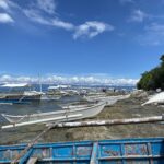 Una fila di bangka bianche ormeggiate lungo una riva rocciosa, piccole piroghe a bilanciere tirate sulla roccia e una barca blu scrostata in primo piano, sotto un cielo azzurro con nuvole bianche.