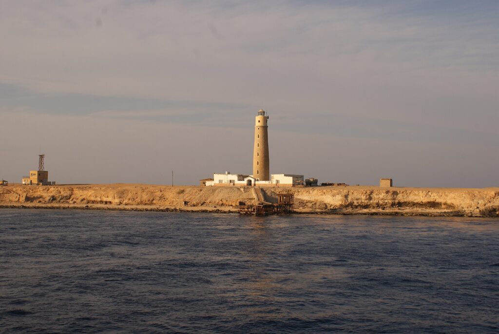 Il faro di Big Brother e i suoi edifici bianchi sull'isolotto desertico, con un pontile arrugginito in primo piano, nella luce dorata della sera