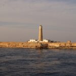 Il faro di Big Brother e i suoi edifici bianchi sull'isolotto desertico, con un pontile arrugginito in primo piano, nella luce dorata della sera