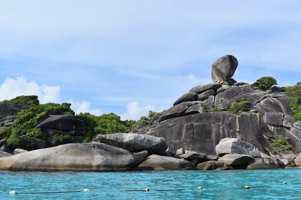 L'enorme masso di granito in equilibrio dell'isola 8 sovrasta un ammasso di rocce grigie e lisce sul bordo di un'acqua turchese, sotto un cielo azzurro velato, con una fila di boe in primo piano