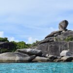 L'enorme masso di granito in equilibrio dell'isola 8 sovrasta un ammasso di rocce grigie e lisce sul bordo di un'acqua turchese, sotto un cielo azzurro velato, con una fila di boe in primo piano