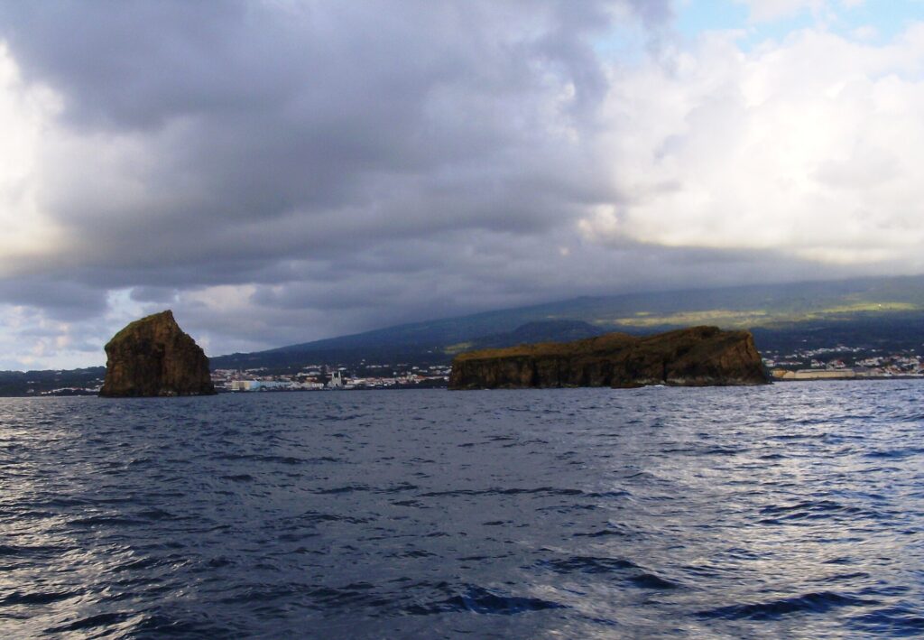 Visti da una barca su un mare scuro e mosso, un isolotto roccioso alto a sinistra e un isolotto lungo e piatto a destra, davanti alla città di Madalena e ai pendii del Pico sotto un cielo carico