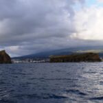 Visti da una barca su un mare scuro e mosso, un isolotto roccioso alto a sinistra e un isolotto lungo e piatto a destra, davanti alla città di Madalena e ai pendii del Pico sotto un cielo carico