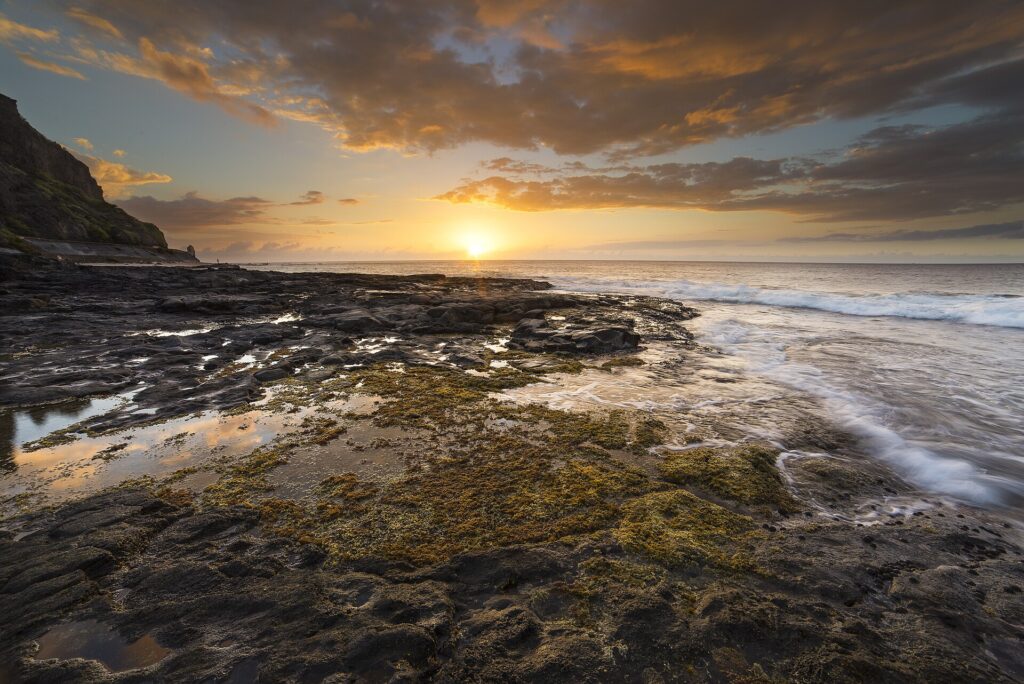 Tramonto sull'oceano visto da una piattaforma di basalto nero coperta di alghe e pozze, con le onde che la lambiscono a destra e una falesia a sinistra