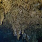Una foresta di stalattiti beige che scende verso l'acqua scura, con una colonna al centro e un bagliore verde a destra