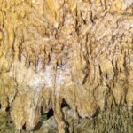 Primo piano di concrezioni giallo pallido e stalattiti sottili, con il riflesso ondulato dell'acqua in basso