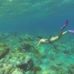 Un'apneista con maschera e pinne rosa scende verso un fondale di rocce e coralli, sotto la superficie increspata, in un'acqua turchese