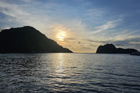 L'isola del Cocco in controluce al tramonto, con una barca da crociera all'ancora sulla destra e il mare calmo