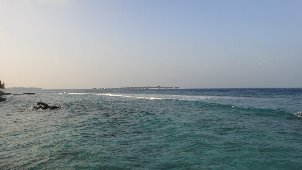 Da una riva rocciosa, un canale turchese e mosso dove una linea di onde si rompe sul reef, e all'orizzonte un'isola bassa orlata di bungalow su palafitte, sotto un cielo pallido.