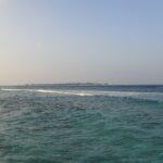 Da una riva rocciosa, un canale turchese e mosso dove una linea di onde si rompe sul reef, e all'orizzonte un'isola bassa orlata di bungalow su palafitte, sotto un cielo pallido.