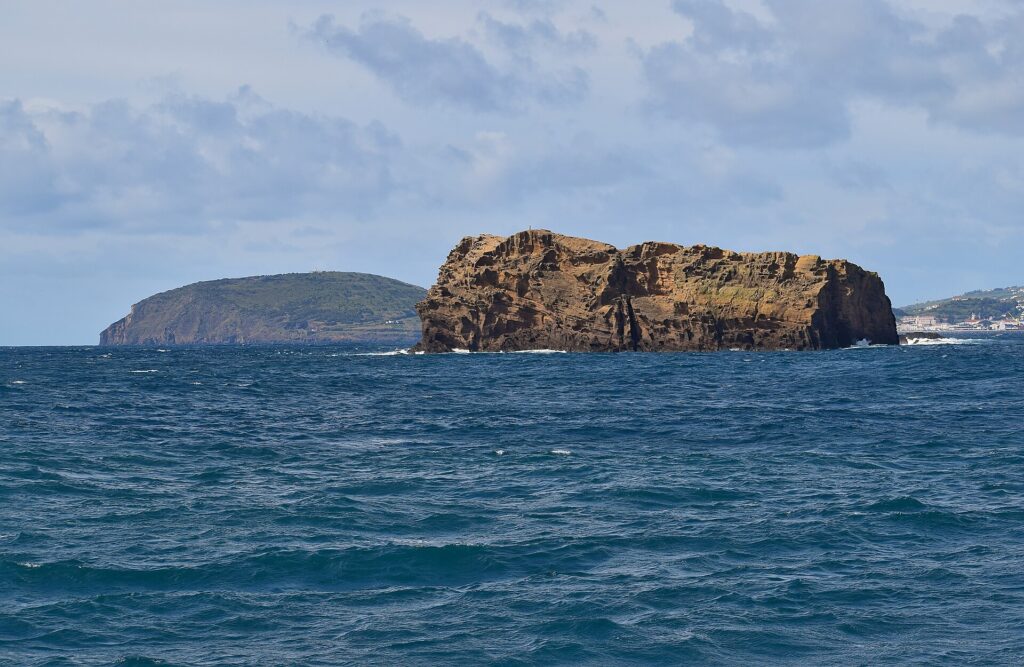 L'Ilhéu Deitado, lungo blocco di tufo ocra con falesie scavate da alveoli, su un mare blu scuro e mosso, con dietro la collina verde del Monte da Guia e le case di Faial in lontananza a destra