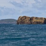 L'Ilhéu Deitado, lungo blocco di tufo ocra con falesie scavate da alveoli, su un mare blu scuro e mosso, con dietro la collina verde del Monte da Guia e le case di Faial in lontananza a destra
