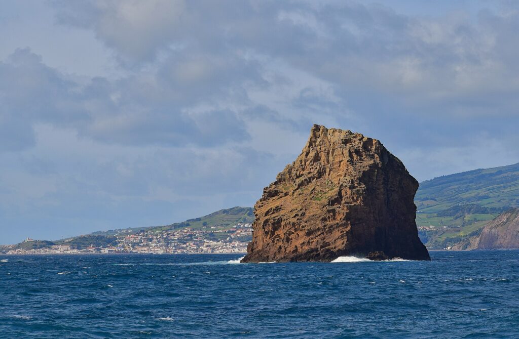L'Ilhéu em Pé, alto scoglio di tufo ocra dagli strati scavati, si erge su un mare blu scuro dove si rompe qualche onda, davanti alla città bianca di Horta e alle colline verdi di Faial sotto un cielo nuvoloso