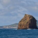 L'Ilhéu em Pé, alto scoglio di tufo ocra dagli strati scavati, si erge su un mare blu scuro dove si rompe qualche onda, davanti alla città bianca di Horta e alle colline verdi di Faial sotto un cielo nuvoloso