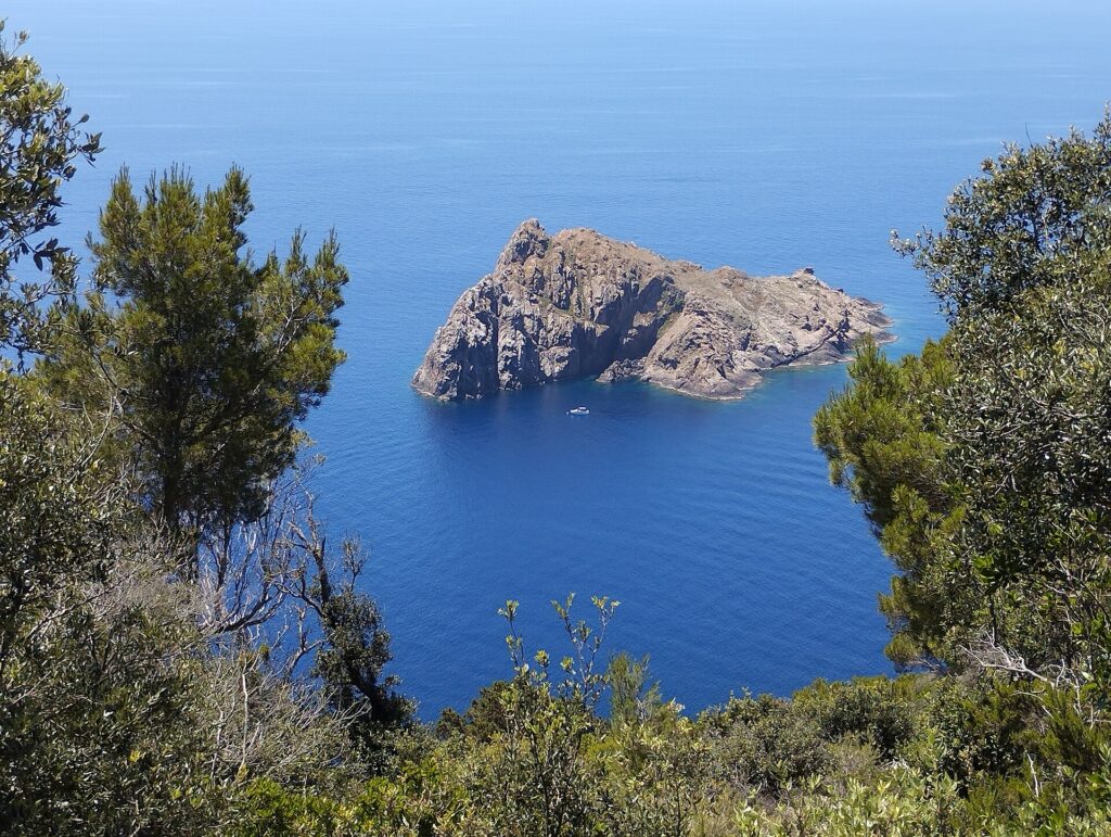 Vista dall'alto di Port-Cros, tra pini e arbusti, sull'isolotto roccioso e scosceso della Gabinière in un mare blu profondo, con una piccola barca bianca ormeggiata ai suoi piedi.