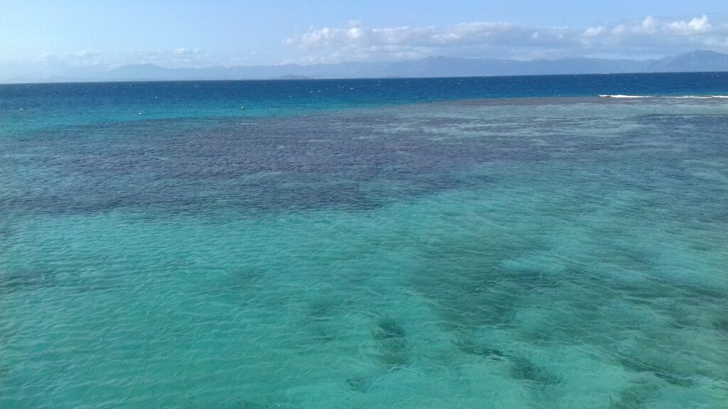 La laguna vista dall'alto, acqua turchese punteggiata di formazioni coralline scure, a destra la barriera dove si infrangono le onde e all'orizzonte le montagne della Grande Terre
