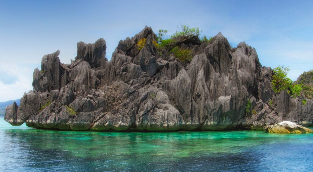 Un isolotto di calcare grigio irto di punte, coperto da un po' di vegetazione, si erge sopra un'acqua turchese trasparente dove si intravede il corallo, sotto un cielo blu.