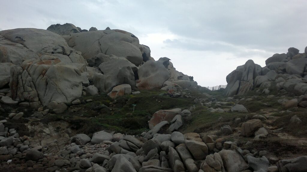 Un ammasso di grandi blocchi di granito grigio arrotondati e scavati dall'erosione, macchiati di licheni arancioni, intorno a un prato raso dove un sentiero delimitato da una staccionata di legno corre verso il mare, sotto un cielo coperto.