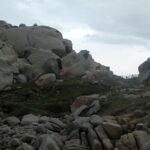 Un ammasso di grandi blocchi di granito grigio arrotondati e scavati dall'erosione, macchiati di licheni arancioni, intorno a un prato raso dove un sentiero delimitato da una staccionata di legno corre verso il mare, sotto un cielo coperto.