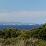 Vista dalla macchia della costa su una lunga isola bassa fatta di blocchi di granito chiaro, in un mare blu scuro, con una barca a vela ai suoi piedi e montagne azzurrine all'orizzonte.