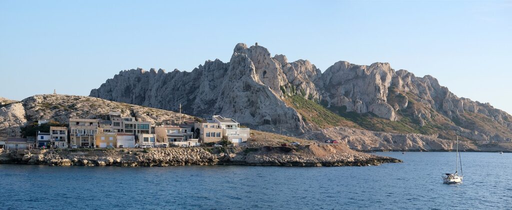 Le case del villaggio di Les Goudes allineate a filo di una costa rocciosa, davanti a un'alta cresta di calcare bianco dalle punte frastagliate, sotto un cielo blu, con una barca a vela all'ancora sul mare calmo.