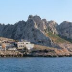 Le case del villaggio di Les Goudes allineate a filo di una costa rocciosa, davanti a un'alta cresta di calcare bianco dalle punte frastagliate, sotto un cielo blu, con una barca a vela all'ancora sul mare calmo.