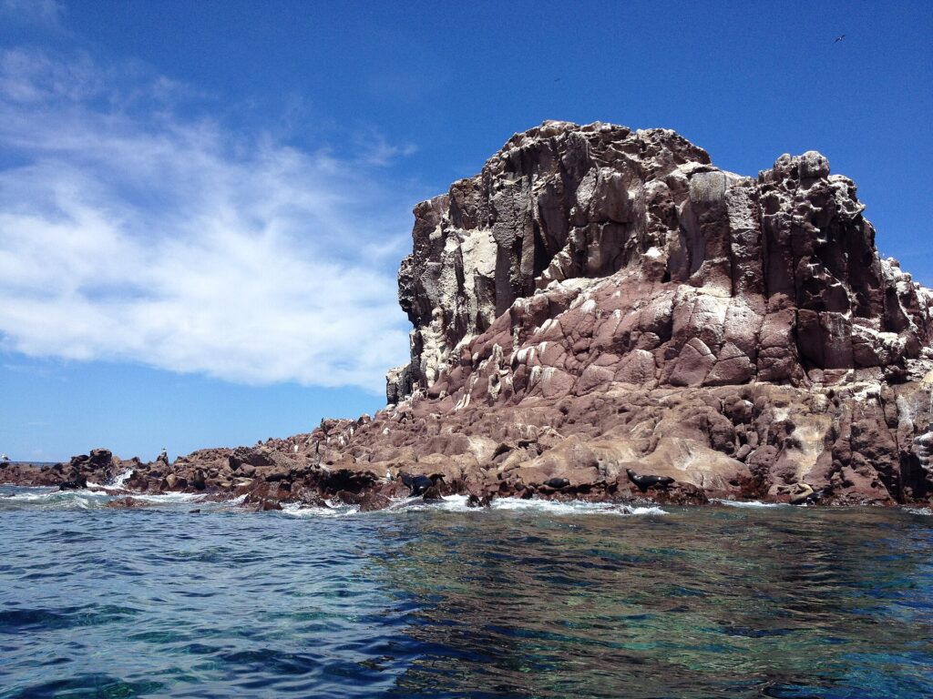 Le rocce rossicce di Los Islotes sotto un cielo blu, con leoni marini sdraiati a filo d'acqua e una scogliera crepata sopra