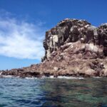 Le rocce rossicce di Los Islotes sotto un cielo blu, con leoni marini sdraiati a filo d'acqua e una scogliera crepata sopra