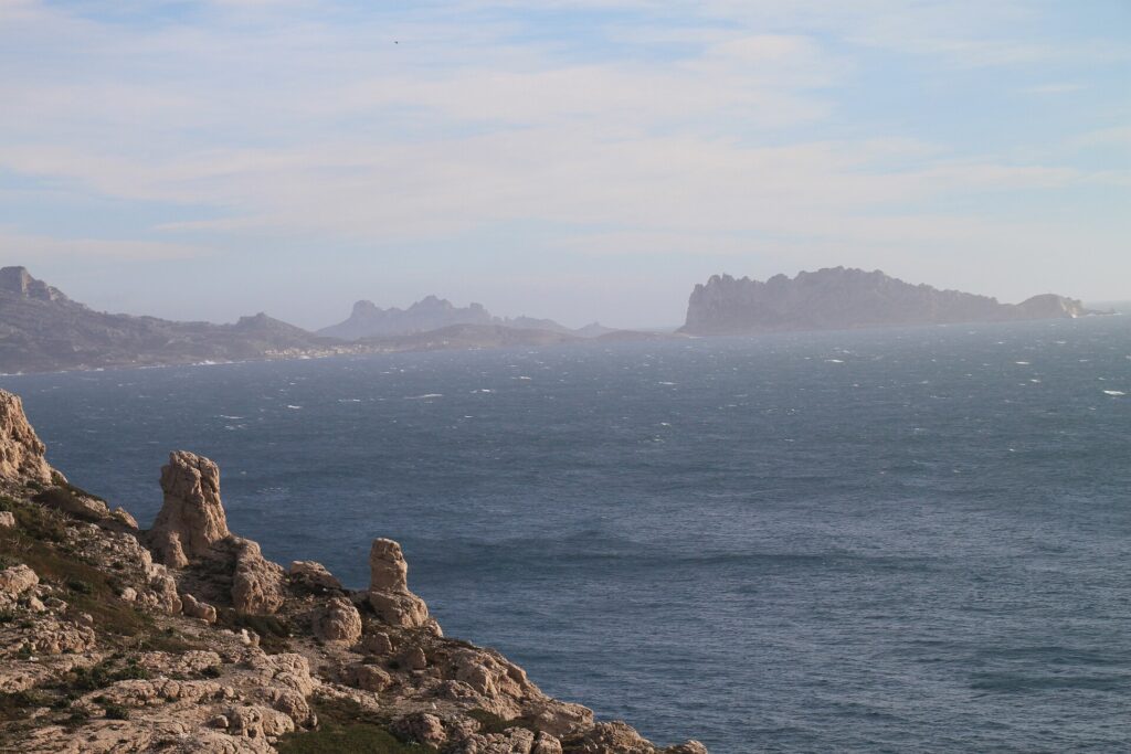 Dalle rocce calcaree del Frioul in primo piano, vista su un mare agitato e spumeggiante fino all'isola Maïre e alla costa di Les Goudes, nella foschia.