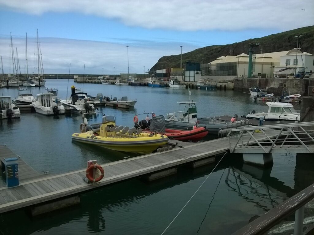 Gommoni, uno giallo con file di sedili e uno rosso, ormeggiati al pontile del porto turistico di Vila do Porto, barche a vela e pescherecci sullo sfondo sotto una scogliera verde e un cielo nuvoloso