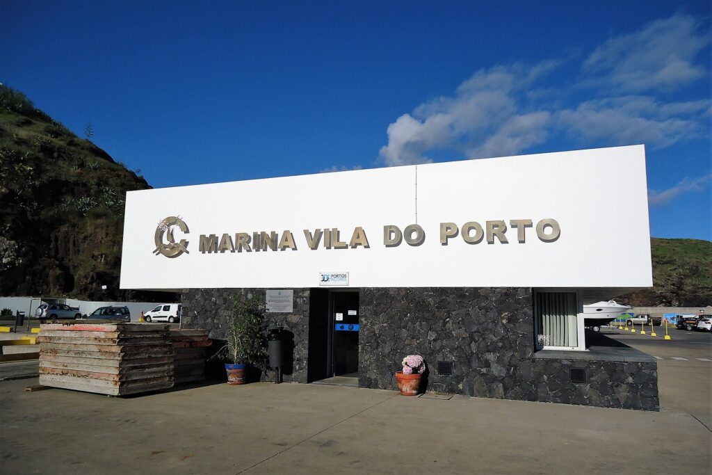 L'edificio di accoglienza del porto turistico di Vila do Porto, grande insegna bianca a lettere metalliche su un muro di basalto nero, sotto un cielo azzurro, una scogliera verde a sinistra