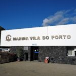 L'edificio di accoglienza del porto turistico di Vila do Porto, grande insegna bianca a lettere metalliche su un muro di basalto nero, sotto un cielo azzurro, una scogliera verde a sinistra