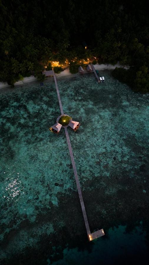 Vista aerea notturna di un lungo pontile in legno che parte dalla spiaggia, con due capanne su palafitte a metà strada
