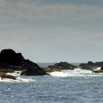 Panorama della catena di scogli neri delle Formigas a filo d'acqua, orlati di schiuma bianca su un mare blu scintillante, con il faro bianco di Formigão a destra sotto un cielo velato