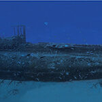 Panoramica subacquea di tutta la lunghezza del relitto del Rubis, adagiato dritto sulla sabbia, con la torretta al centro, in un'acqua blu punteggiata di pesci.