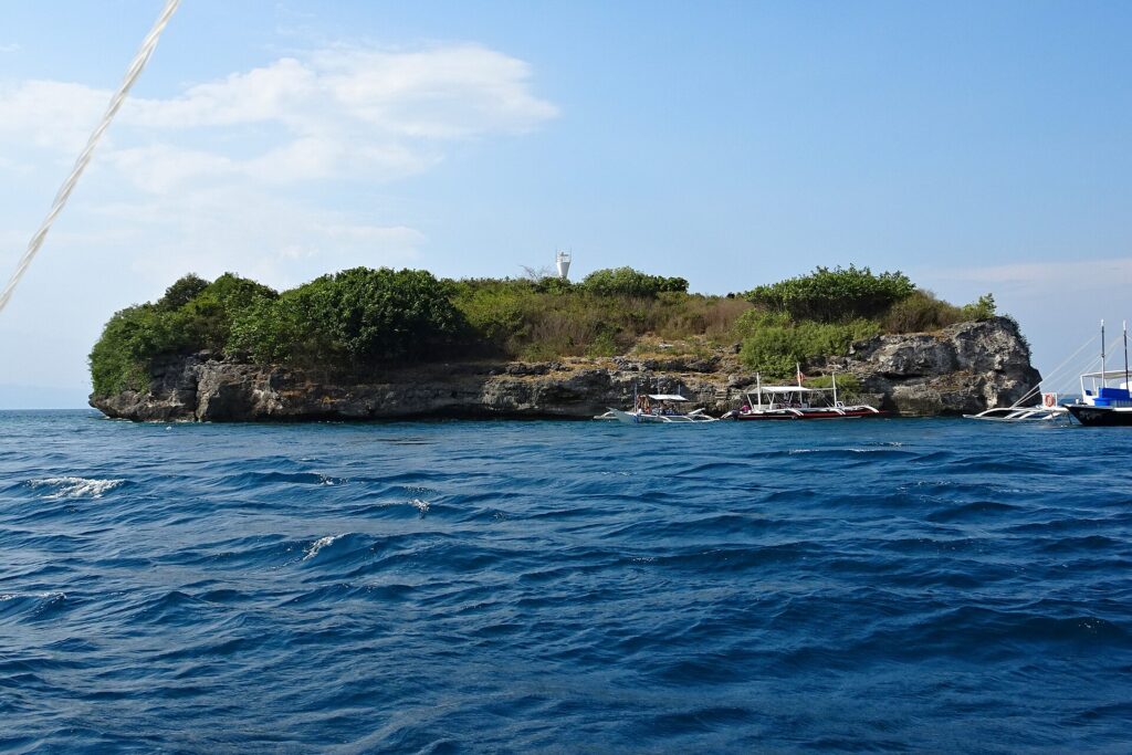 L'isola di Pescador vista dal mare, coperta di cespugli verdi, con il faro bianco in cima e tre banca da immersione ormeggiate ai piedi della falesia