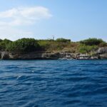 L'isola di Pescador vista dal mare, coperta di cespugli verdi, con il faro bianco in cima e tre banca da immersione ormeggiate ai piedi della falesia