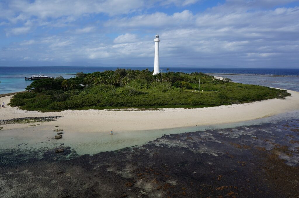 Il grande faro bianco dell'isolotto Amédée svetta sopra la vegetazione, l'isolotto è orlato di sabbia bianca, in primo piano la piattaforma corallina scoperta e a sinistra una barca all'ormeggio