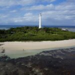 Il grande faro bianco dell'isolotto Amédée svetta sopra la vegetazione, l'isolotto è orlato di sabbia bianca, in primo piano la piattaforma corallina scoperta e a sinistra una barca all'ormeggio