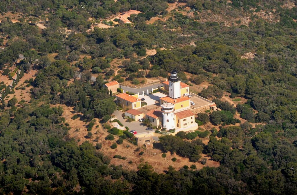 Vista aerea del faro di capo Camarat, torre bianca con lanterna nera circondata da edifici gialli dai tetti di tegole arancioni, in mezzo a una fitta macchia mediterranea.