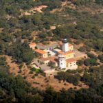 Vista aerea del faro di capo Camarat, torre bianca con lanterna nera circondata da edifici gialli dai tetti di tegole arancioni, in mezzo a una fitta macchia mediterranea.