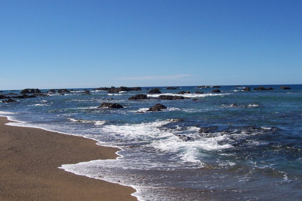 Una spiaggia di sabbia bruno dorata dove muoiono onde orlate di schiuma, davanti a un mare blu punteggiato di rocce nere fino all'orizzonte, sotto un cielo senza nuvole.