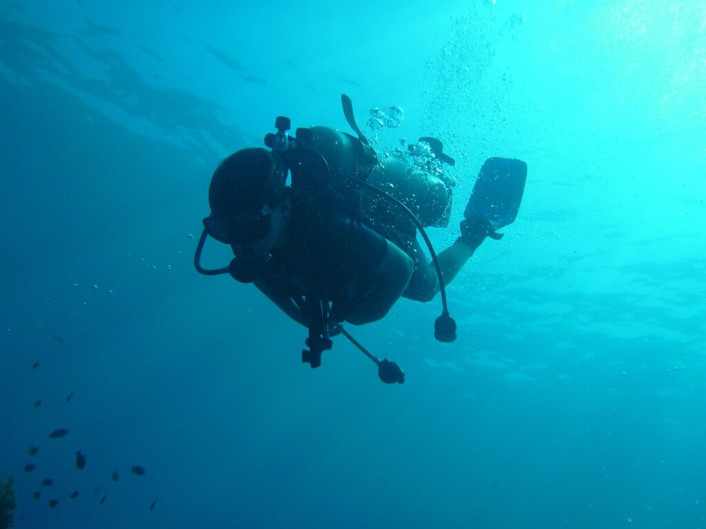 Un sub in silhouette visto dal basso, con bombola, erogatori e pinne stagliati sull'acqua turchese, e le bolle che salgono verso la superficie illuminata.