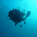 Un sub in silhouette visto dal basso, con bombola, erogatori e pinne stagliati sull'acqua turchese, e le bolle che salgono verso la superficie illuminata.