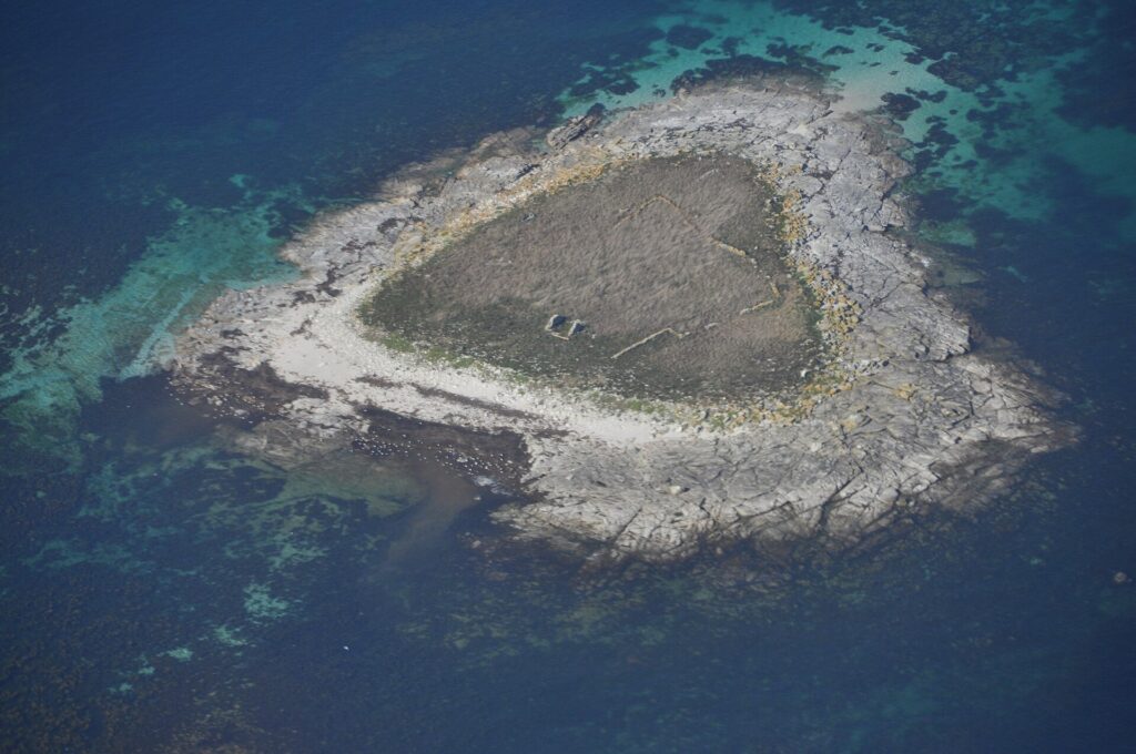 Vista aerea di un piccolo isolotto di lastre di granito grigio, coperto al centro di erba secca dove si intuiscono muretti in rovina, orlato da una spiaggia chiara e circondato da bassifondi turchesi e acqua blu profondo.