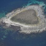 Vista aerea di un piccolo isolotto di lastre di granito grigio, coperto al centro di erba secca dove si intuiscono muretti in rovina, orlato da una spiaggia chiara e circondato da bassifondi turchesi e acqua blu profondo.