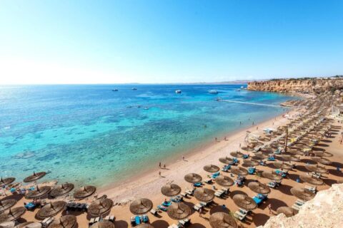 Una lunga spiaggia di sabbia ai piedi di una scogliera, con ombrelloni di paglia e un pontile sul mare turchese
