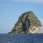 Il Rocher du Diamant visto da vicino dal mare, una massa di roccia grigia scavata da grotte, striata di bianco e coperta in cima di vegetazione, sopra un mare blu scuro